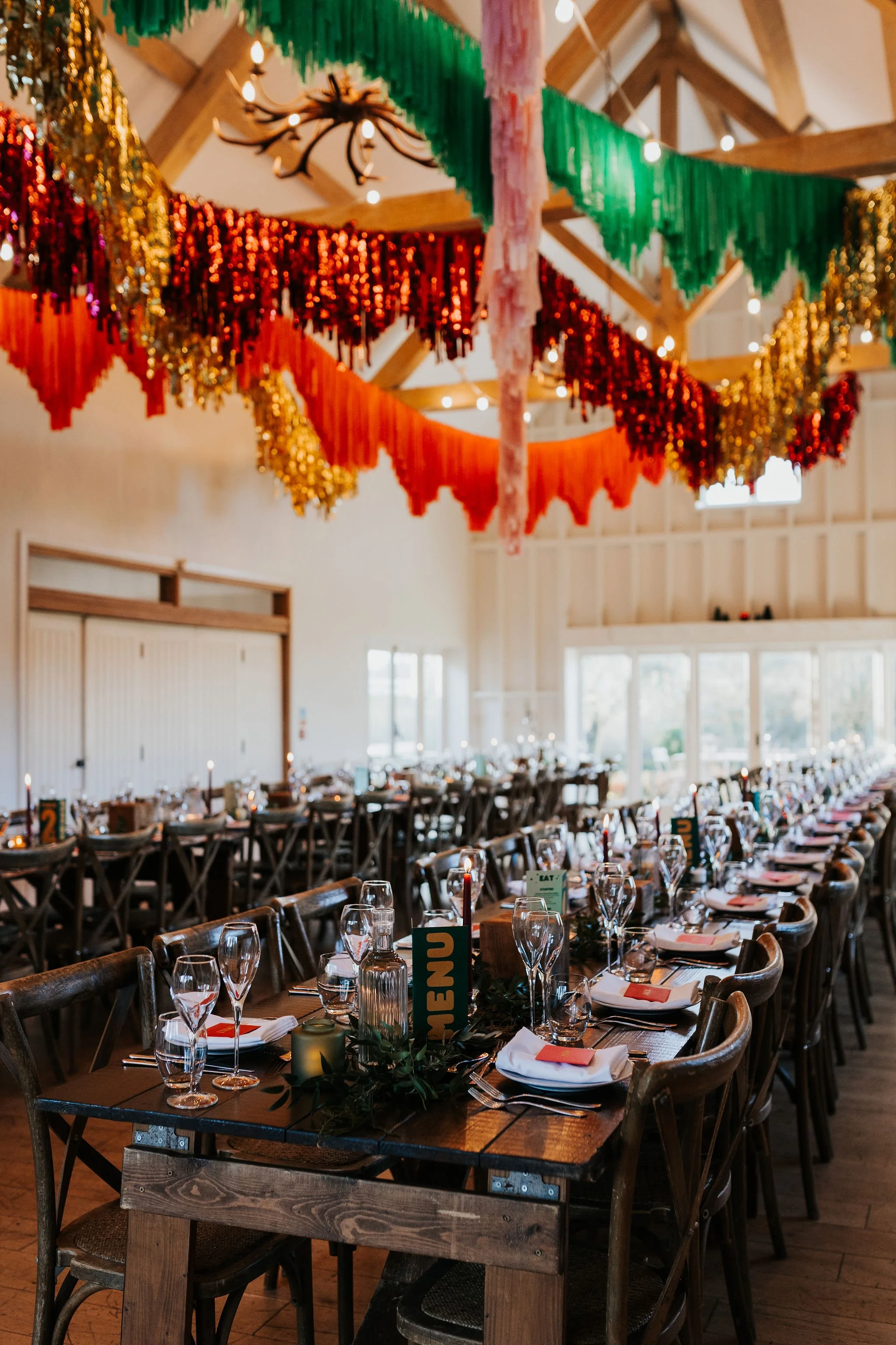 Modern wedding reception by AMW Events, featuring long wooden banquet tables with streamer decorations hanging from exposed beams. SJ Wed Photo.