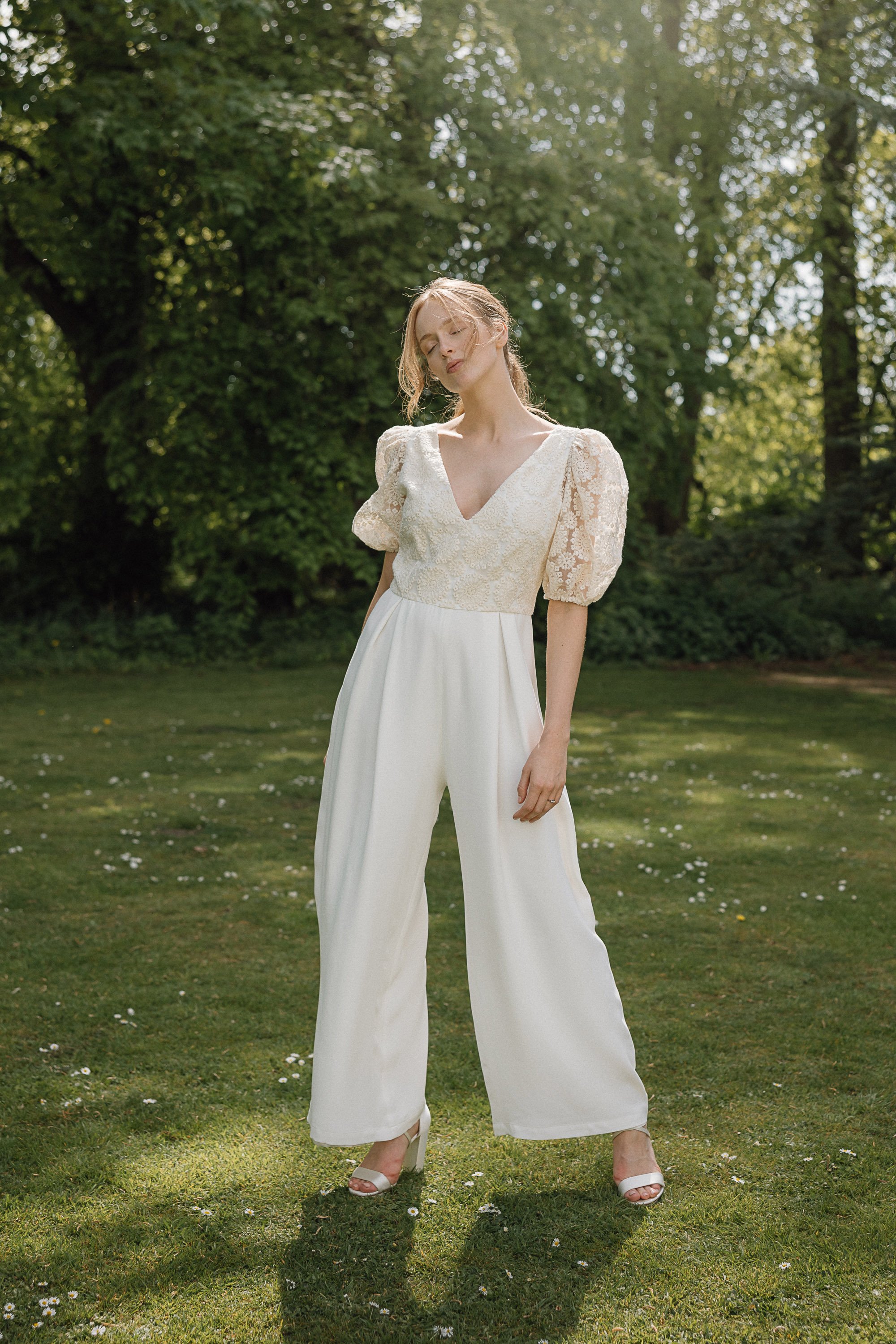 The bride stands on sunlit grass surrounded by soft greenery wearing an elegant ivory jumpsuit with gently flared trousers and a deep V neckline, the bodice crafted from delicate floral lace with subtle texture and short puffed sleeves that add a rom