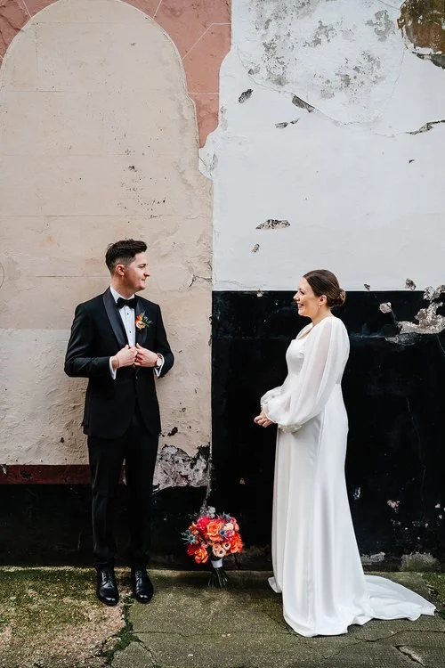 A couple stand facing one another against a textured, weathered wall that blends soft pastels with peeling paint, creating an artistic backdrop for their moment together. The groom adjusts his suit jacket while the bride, dressed in an elegant gown w