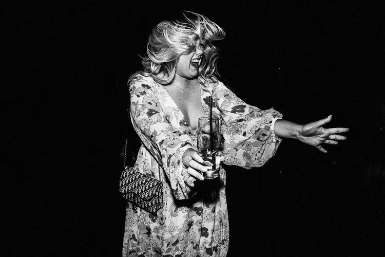 A high-energy black and white candid in Matt Parry’s signature raw, flash-driven style. A woman dances with total abandon, her hair whipping wildly across her face as she laughs mid-movement. She wears a floral dress with wide, flowing sleeves and ca
