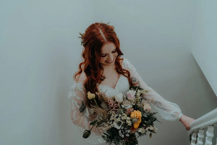 Soft romantic bridal bouquet by New Bud Floristry featuring pastel roses, dried grasses, ranunculus and natural garden foliage. Shown as the bride descends a staircase in an elegant lace gown, this delicate textural arrangement highlights New Bud Flo