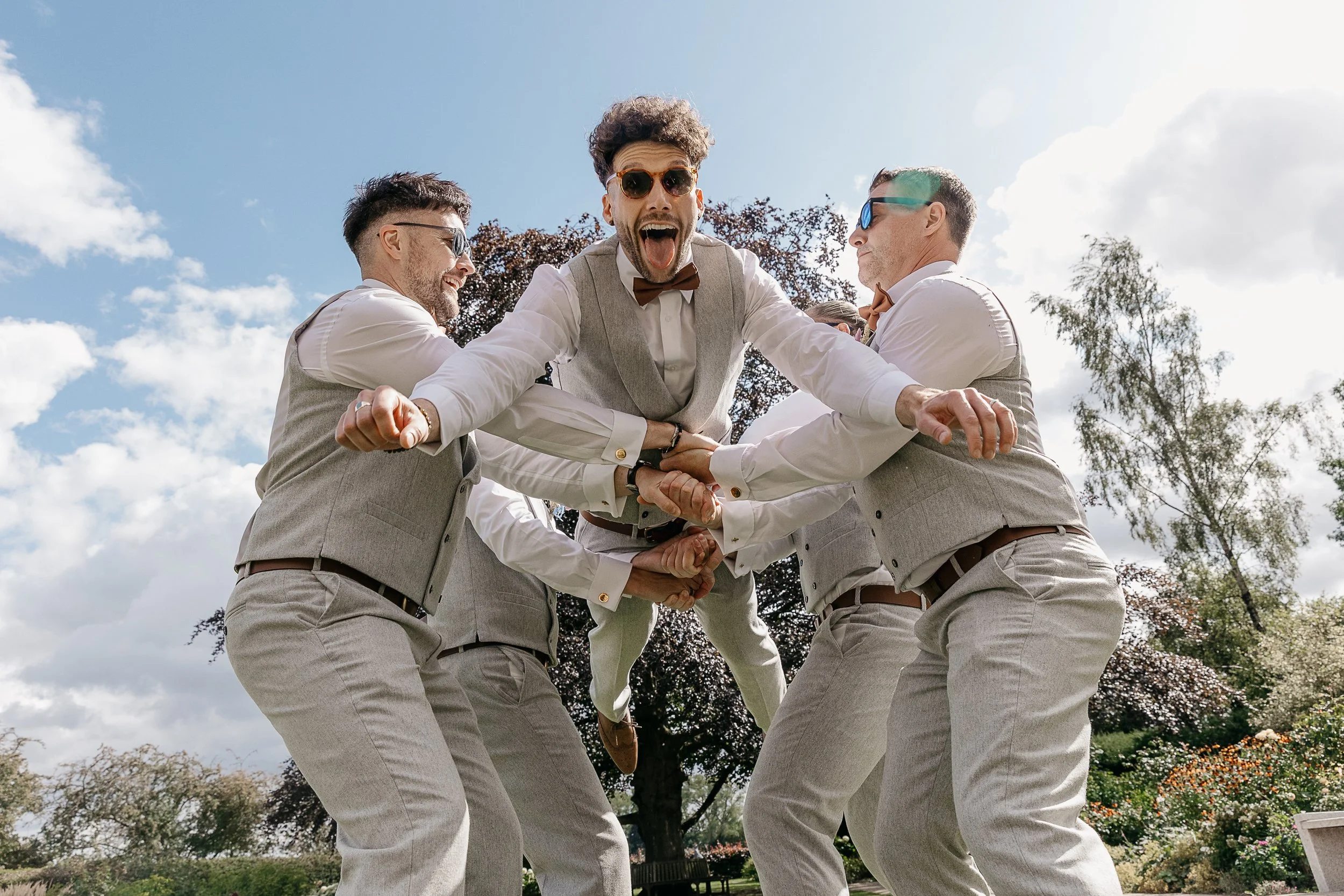 A high energy and brilliantly fun moment captured by YU Photography, showing the groom being launched into the air by his groomsmen as they all burst into laughter. Their matching light grey suits and sunglasses add to the cool, coordinated look whil