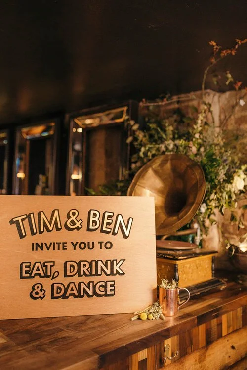 A bespoke handmade wedding sign by Esther of Sylvie Signs featuring the message “Tim & Ben invite you to eat, drink & dance.” The custom wooden sign is painted with bold 3D lettering in black and cream, sitting on a warm wood background. Displayed on