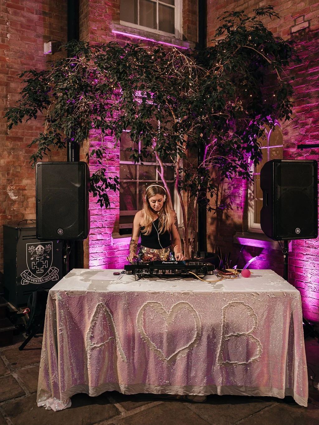 This image shows a lively wedding DJ setup featuring Cara from Rave The Date performing outdoors in front of a warmly lit brick backdrop. She stands behind a sequin-covered table with a heart and the initials “A ♥ B” drawn into the fabric — a playful