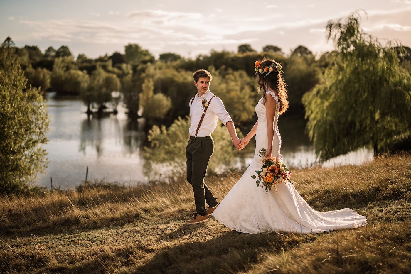 A couple walk hand in hand beside a tranquil lake at sunset, celebrating a nature-led, eco-friendly wedding planned by Wild Soul Weddings, eco wedding planners in the UK. Rach Elizabeth Photography.