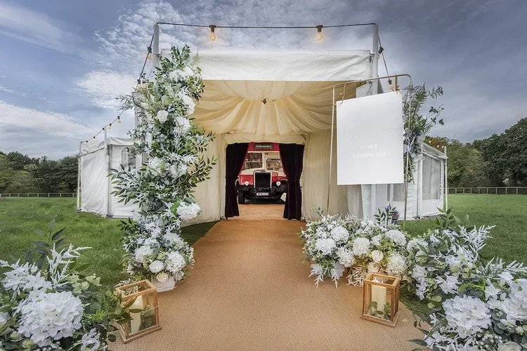 An elegant outdoor wedding setup featuring the Deckerdence Mobile Venue, a luxury double-decker bus bar and marquee. The photo showcases a beautifully styled marquee entrance lined with lush white floral arrangements, gold lanterns, and soft draped f