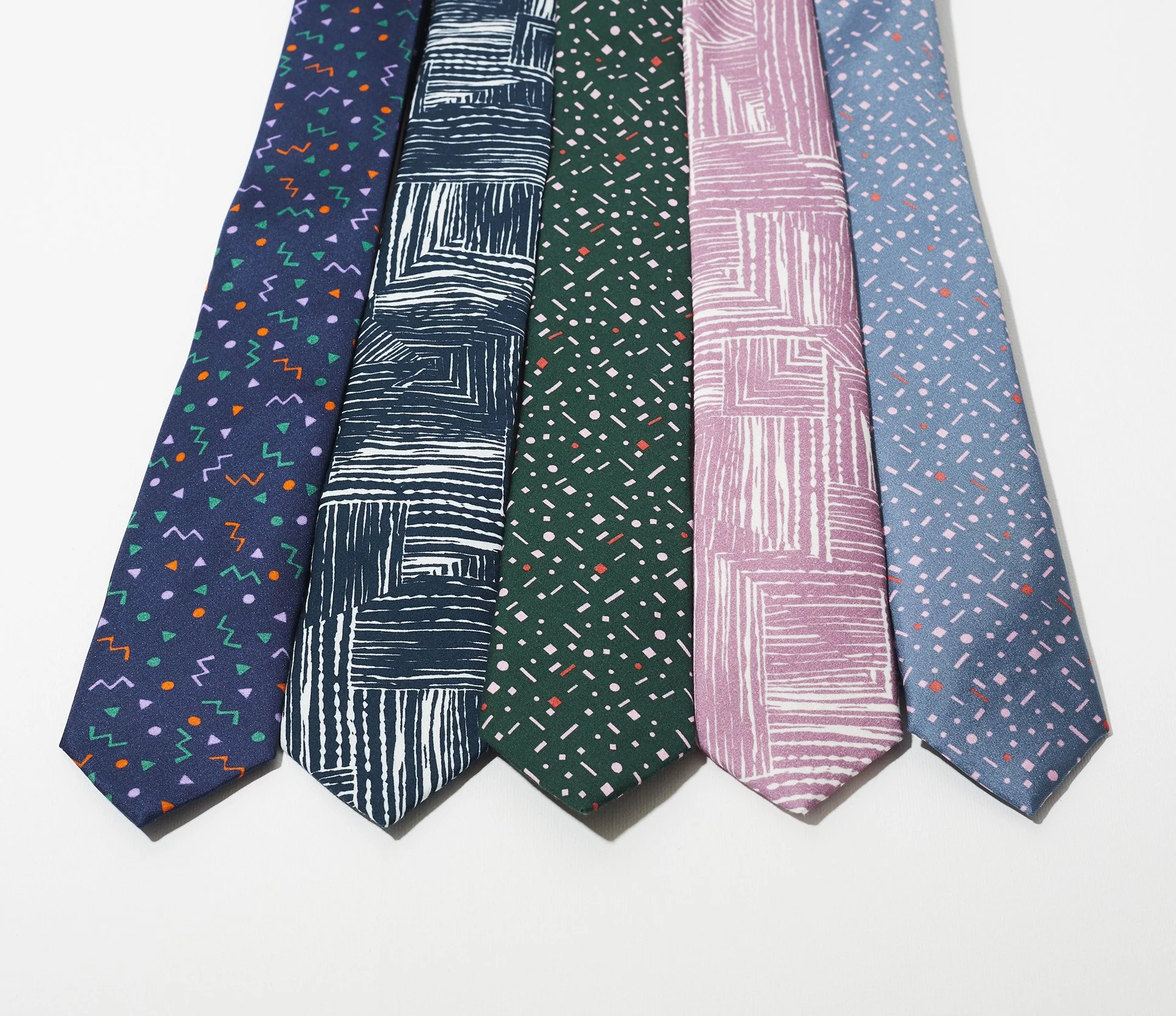 A row of patterned Francis + Louise ties is arranged side by side on a white surface, featuring a navy design with playful multicoloured shapes, a dark blue tie with bold linear blocks, a deep green tie scattered with small geometric marks, a soft ma