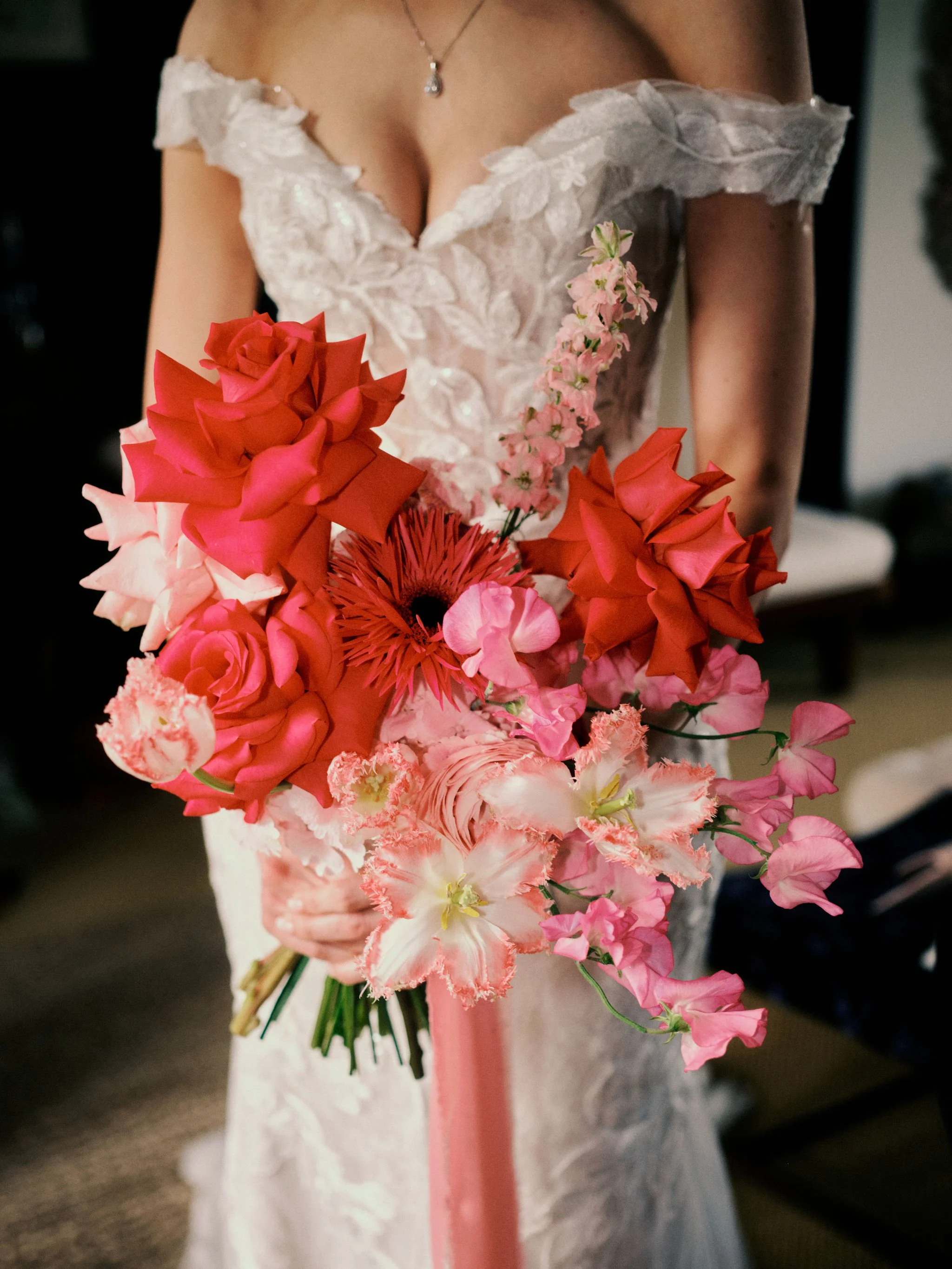 Romantic bridal bouquet by Sweet Pea Sorbet Floristry featuring bold red and pink roses, sweet peas, and frilled tulips. Captured by Ochi Weddings, the vibrant arrangement complements the bride’s lace off-shoulder gown beautifully.