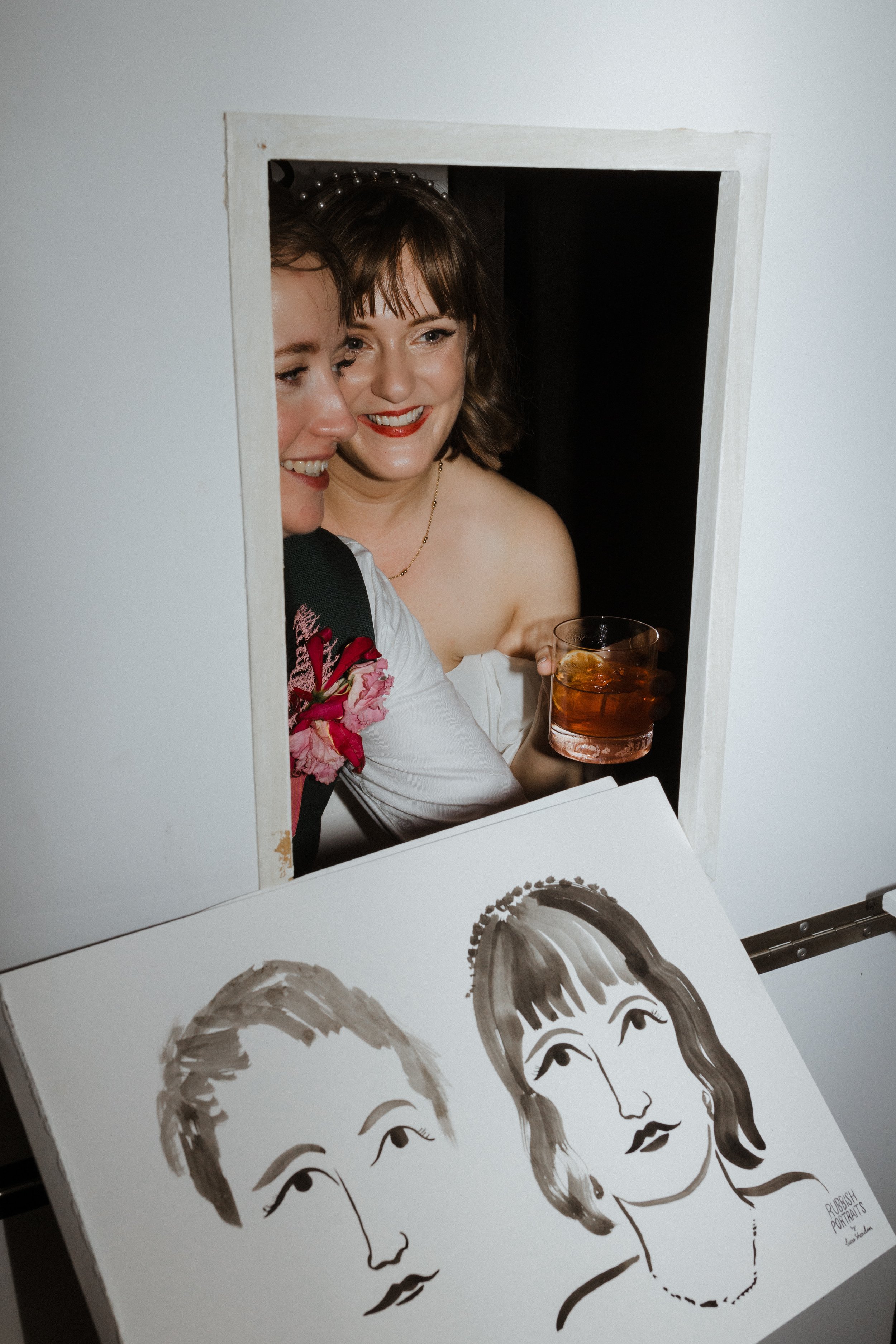 A joyful moment inside the Rubbish Portraits booth as two guests peek through the portrait window, smiling while admiring their freshly painted artwork. The ink illustration captures their likeness in Lucie Sheridan’s signature playful style, making 