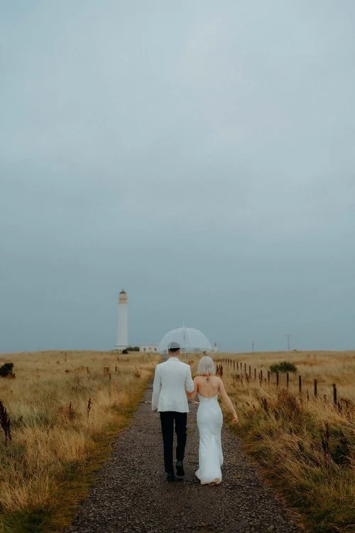 A beautifully atmospheric moment captured by F.D Young Photography: a couple walking hand-in-hand down a rugged coastal path beneath a soft, moody sky. Sharing a clear umbrella and framed by wild grasses and a distant lighthouse, they move together w