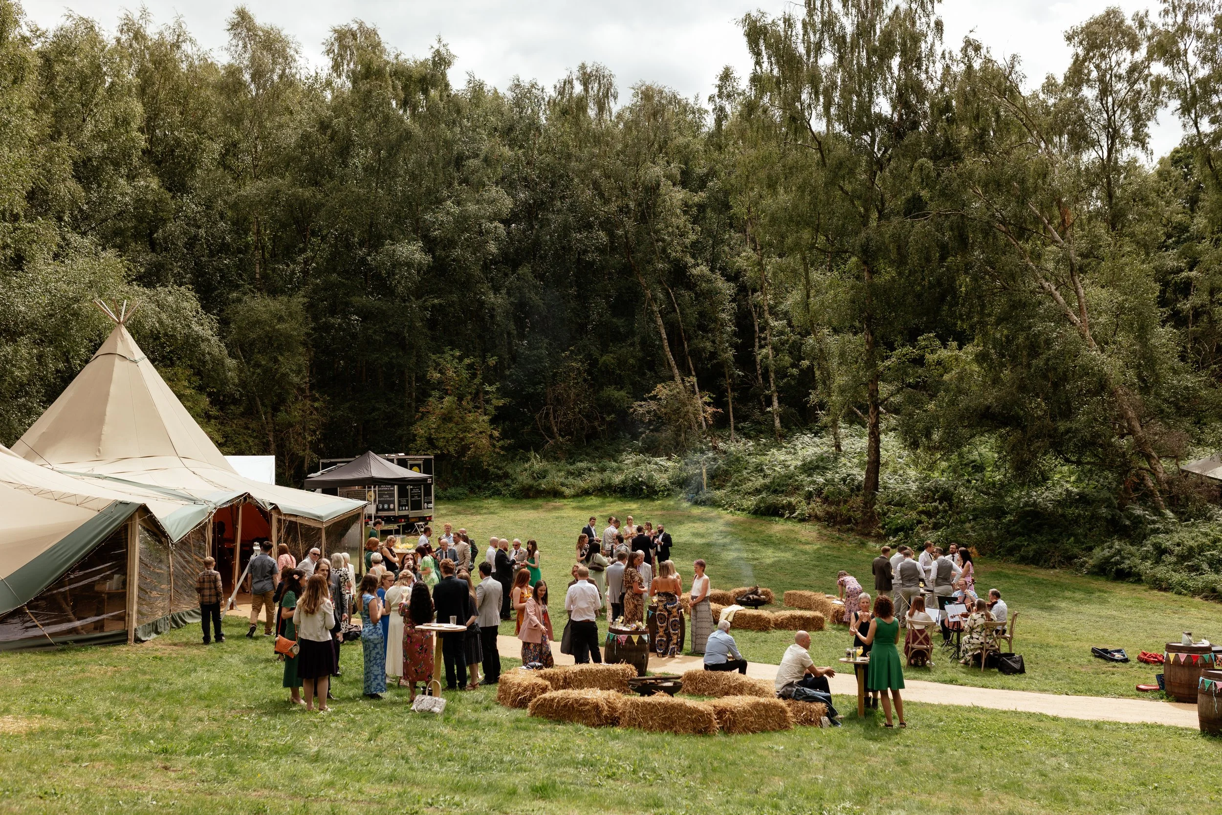 An outdoor woodland wedding at Sherwood Glade. There are trees, guests, tipis and hay bails.