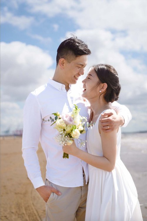 A couple share a warm embrace on a sunlit sandy beach, smiling at one another as they hold a soft pastel bouquet, beautifully captured by Christy Photography. The bright sky, gentle coastal breeze and natural light create a relaxed and romantic atmos