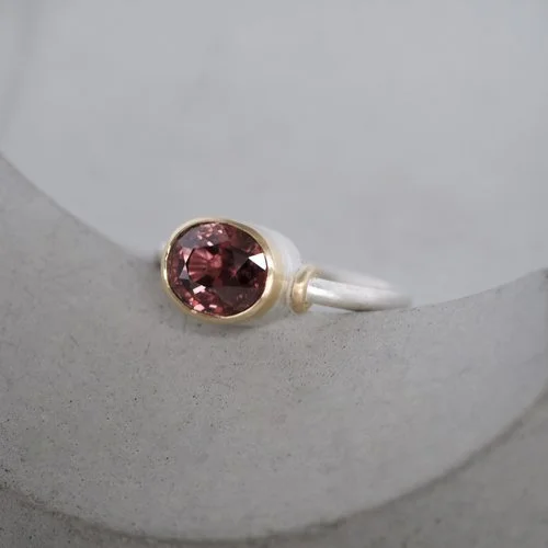 A handcrafted Natalie Vernazza engagement ring featuring a rich pink Malay zircon set in an 18ct yellow gold bezel on a softly textured Argentium silver band, photographed on a minimalist stone backdrop to highlight its organic, contemporary wedding 