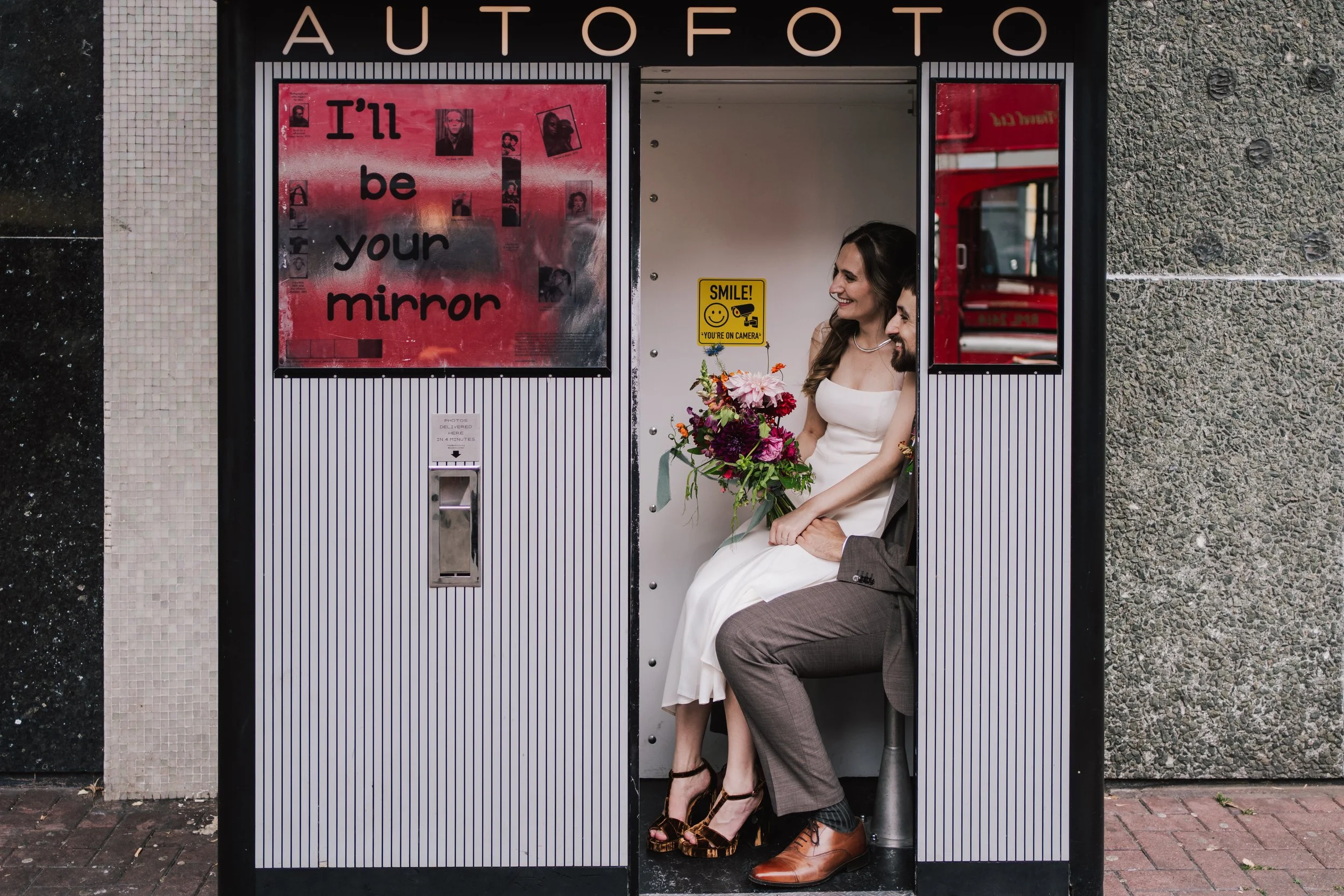 A couple squeeze into a retro street photo booth, laughing together as they sit side by side with the bride’s vibrant bouquet resting in her lap. The bold “I’ll be your mirror” message displayed on the booth’s exterior adds a playful, artistic touch,