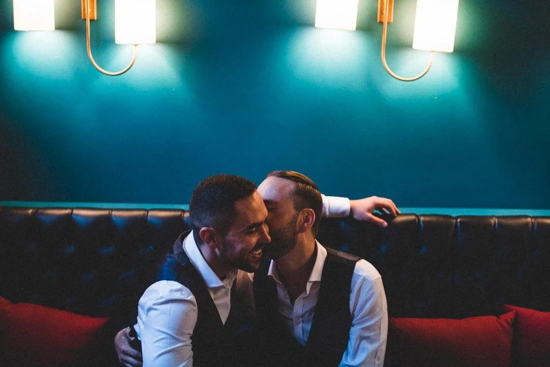 A romantic and atmospheric portrait captured by Barbara K Photography, showing two grooms sharing an affectionate moment in a stylish bar lounge. One kisses the other on the cheek as they sit close together on a dark tufted banquette, lit by the warm