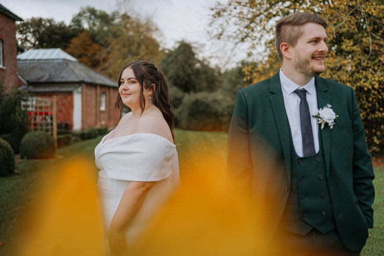 A couple stand back to back in a landscaped garden with soft autumn colours in the foreground, captured by Joshua Humphrey Photography. Their relaxed expressions and elegant outfits create a warm natural wedding portrait set against a charming countr