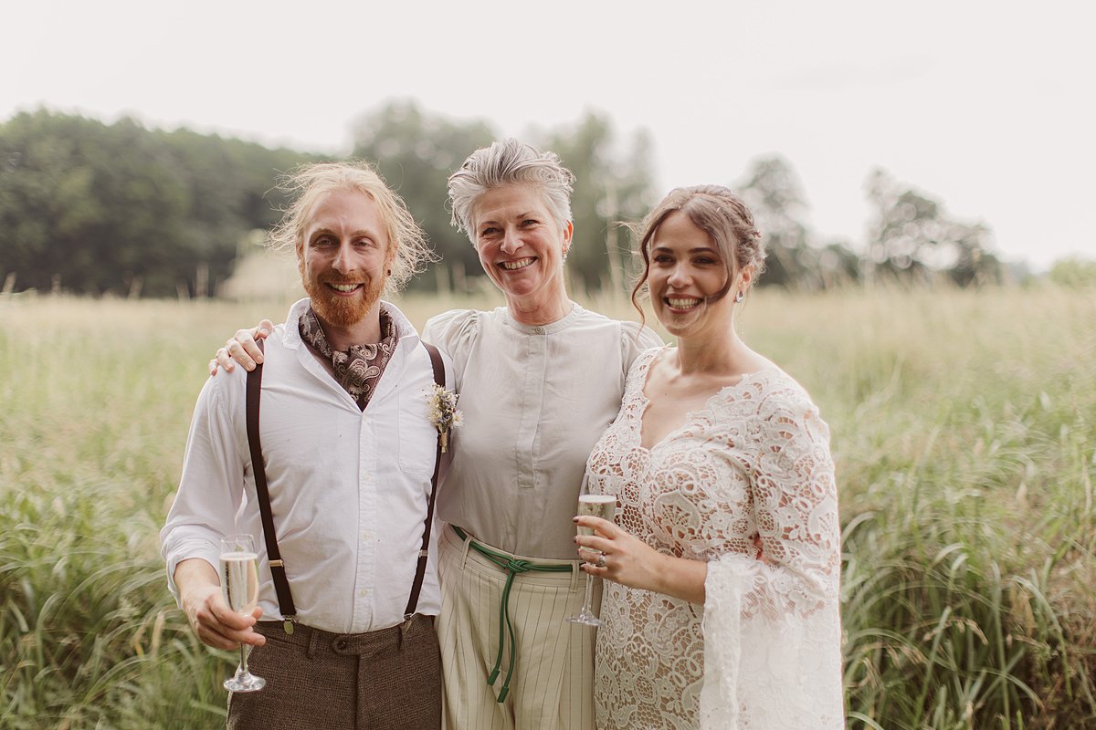 A celebrant stands arm in arm with a couple in a wild meadow, sharing a joyful moment after a heartfelt ceremony led by Arabella of A Wilde Celebrant. Nick Ray Photography.