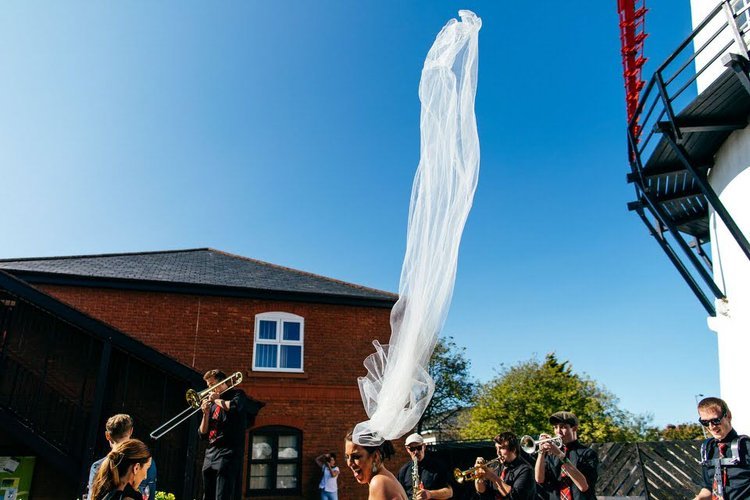 A bride’s veil flies dramatically into the air as a lively brass band performs around her during an outdoor celebration, captured brilliantly by Jordanna Marston Photography. The bright blue sky, movement of the veil and energy of the musicians creat