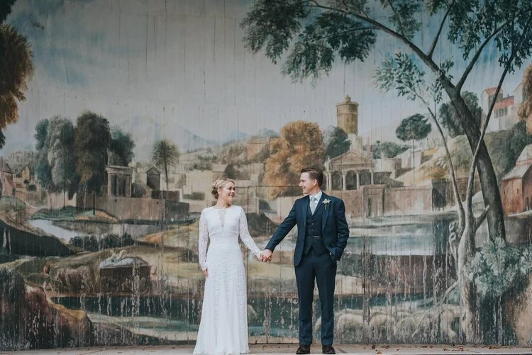 A timeless and elegant wedding portrait captured by Matt Fox Photography, featuring a couple standing hand in hand in front of a large hand painted mural that depicts a serene countryside scene. The bride wears a classic long sleeved lace gown while 