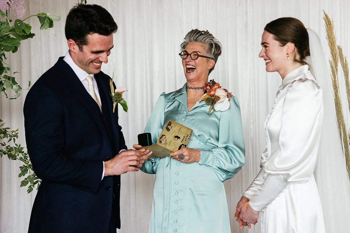 A candid moment during the ceremony Arabella, A Wilde Celebrant, stands between the couple, laughing openly while holding a card, with the couple smiling in response. Lyndsey Goddard Photography.