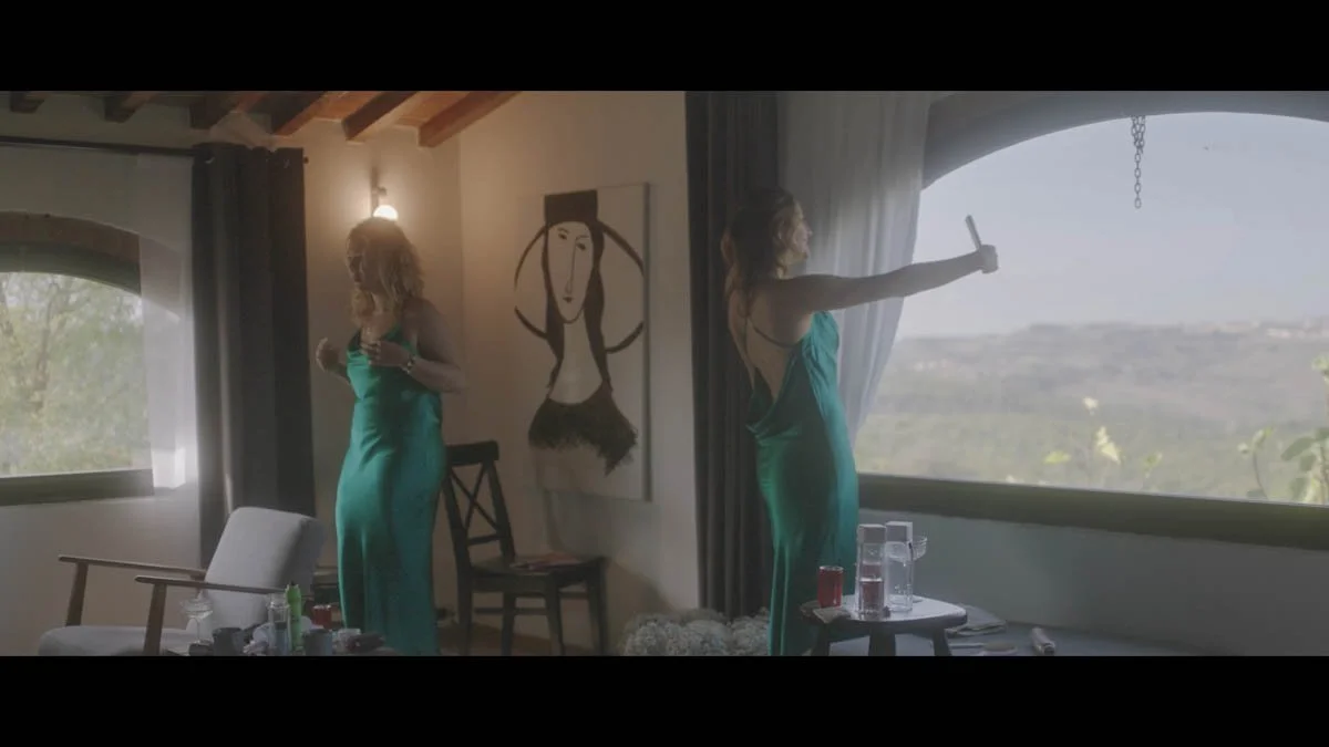 Two bridesmaids in silky emerald dresses move through a softly lit room that overlooks rolling countryside as one admires the view with her phone raised and the other adjusts her outfit creating a serene intimate moment that blends natural beauty wit