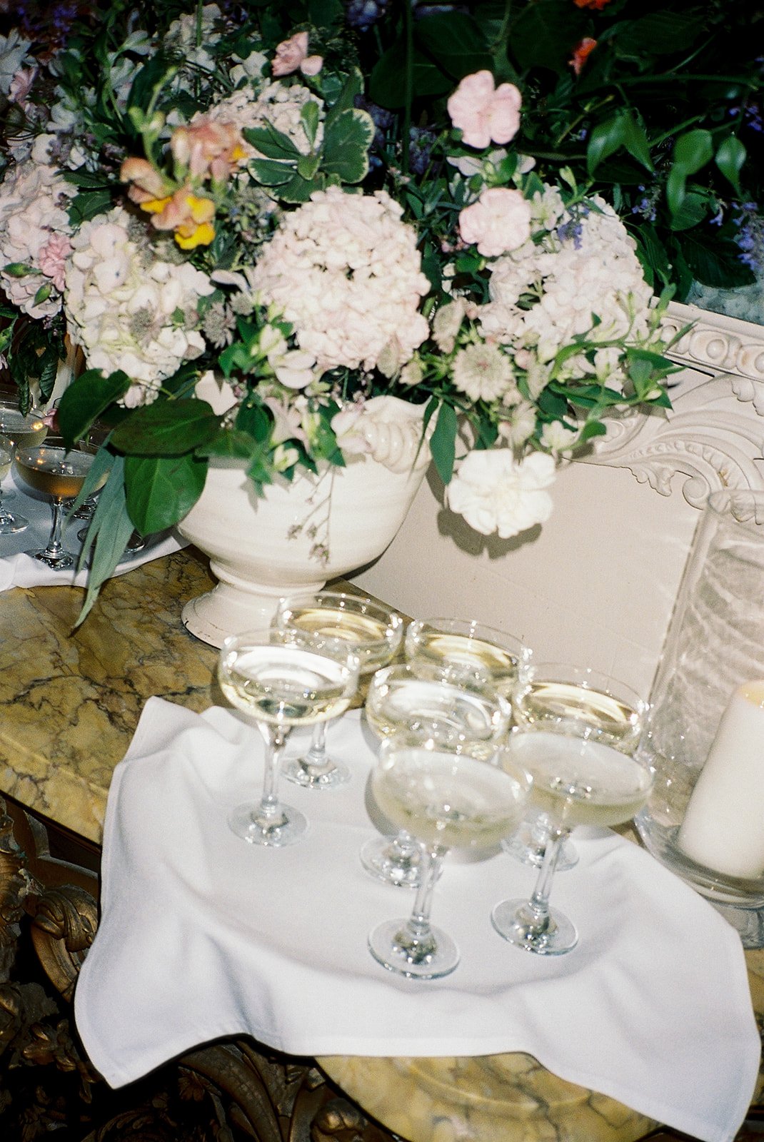 A cluster of coupe glasses filled with pale sparkling wine rests on a white cloth atop a marble table, surrounded by abundant pastel flowers arranged in an elegant urn that adds softness and romance to the refined setting. Tash Busta Photography. 