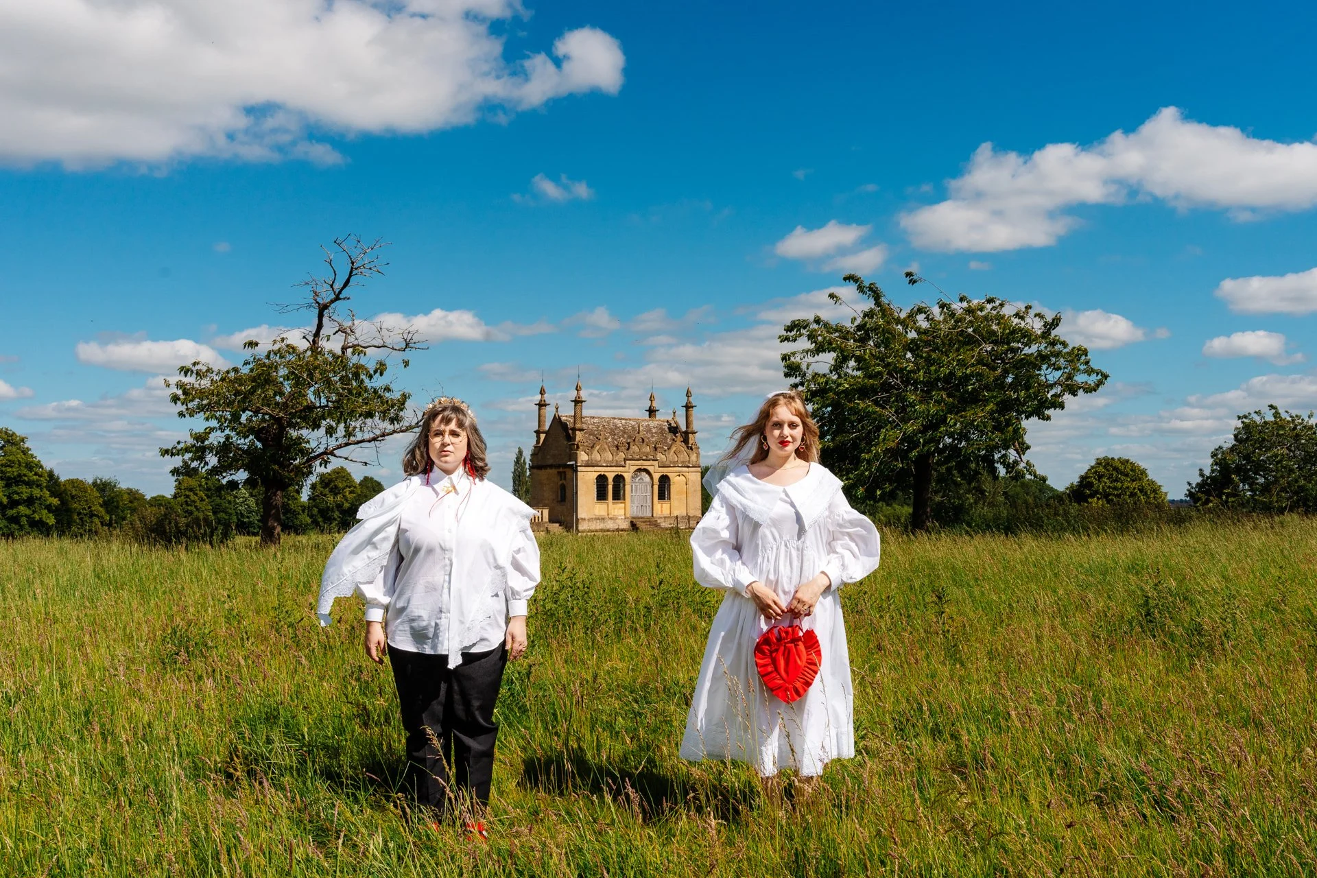 Two people stand in a tall grassy field beneath a bright blue sky, both wearing white outfits, with one holding a red heart-shaped bag. Behind them, an ornate historic stone building rises in the distance, framed by scattered trees and soft clouds. T