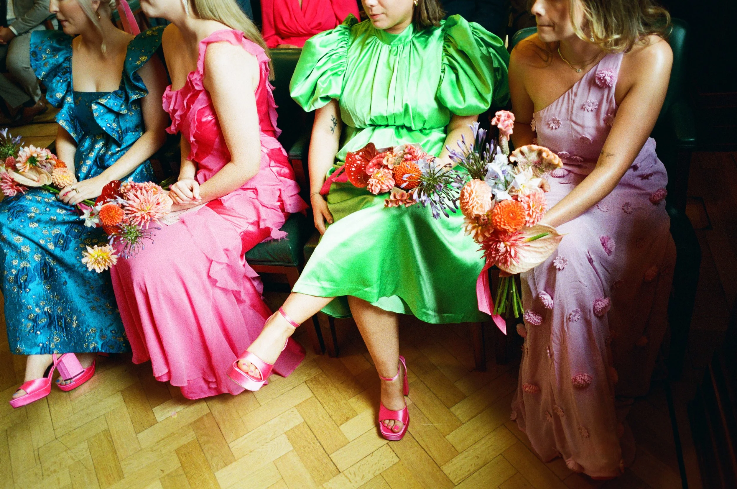 A row of bridesmaids sit together in vibrant, fashion-forward dresses ranging from bold blue brocade to hot pink ruffles, vivid green satin, and soft lilac florals. Each holds a colourful bouquet of textured summer blooms, their bright pink heels add