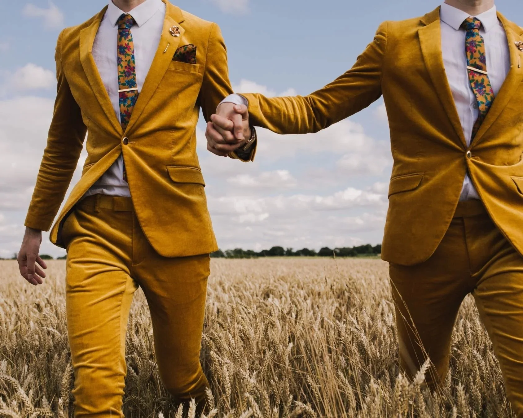 2 grooms in mustard, velver suits are holding hands and walking through a cornfield. Image by Book Of Love Directory member, Lisa Jane Photography