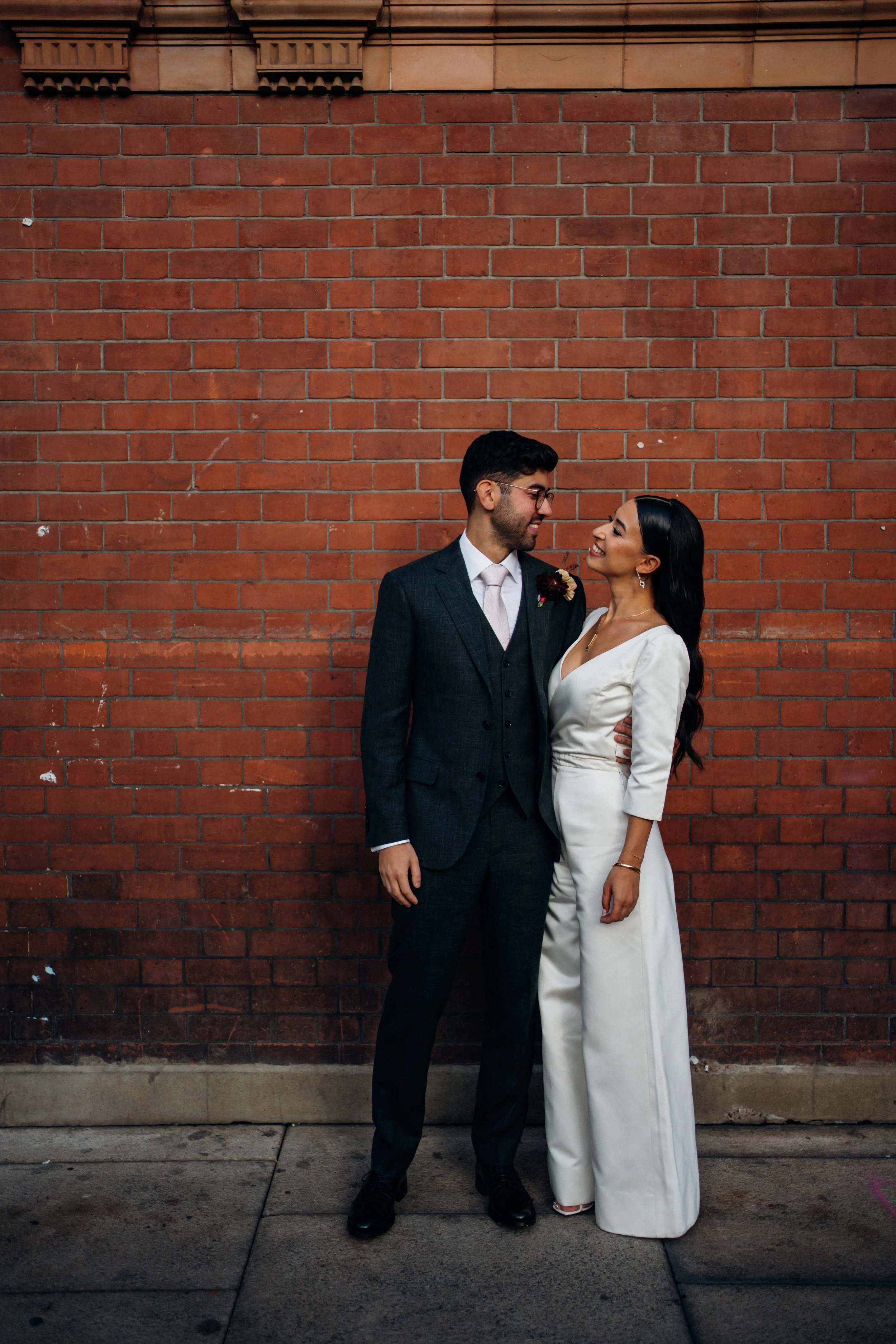 The image shows a couple standing close together on a pavement in front of a red brick wall, looking at each other with warm, happy expressions. The bride is wearing an ivory V neck bridal jumpsuit with three quarter sleeves and wide leg trousers, an