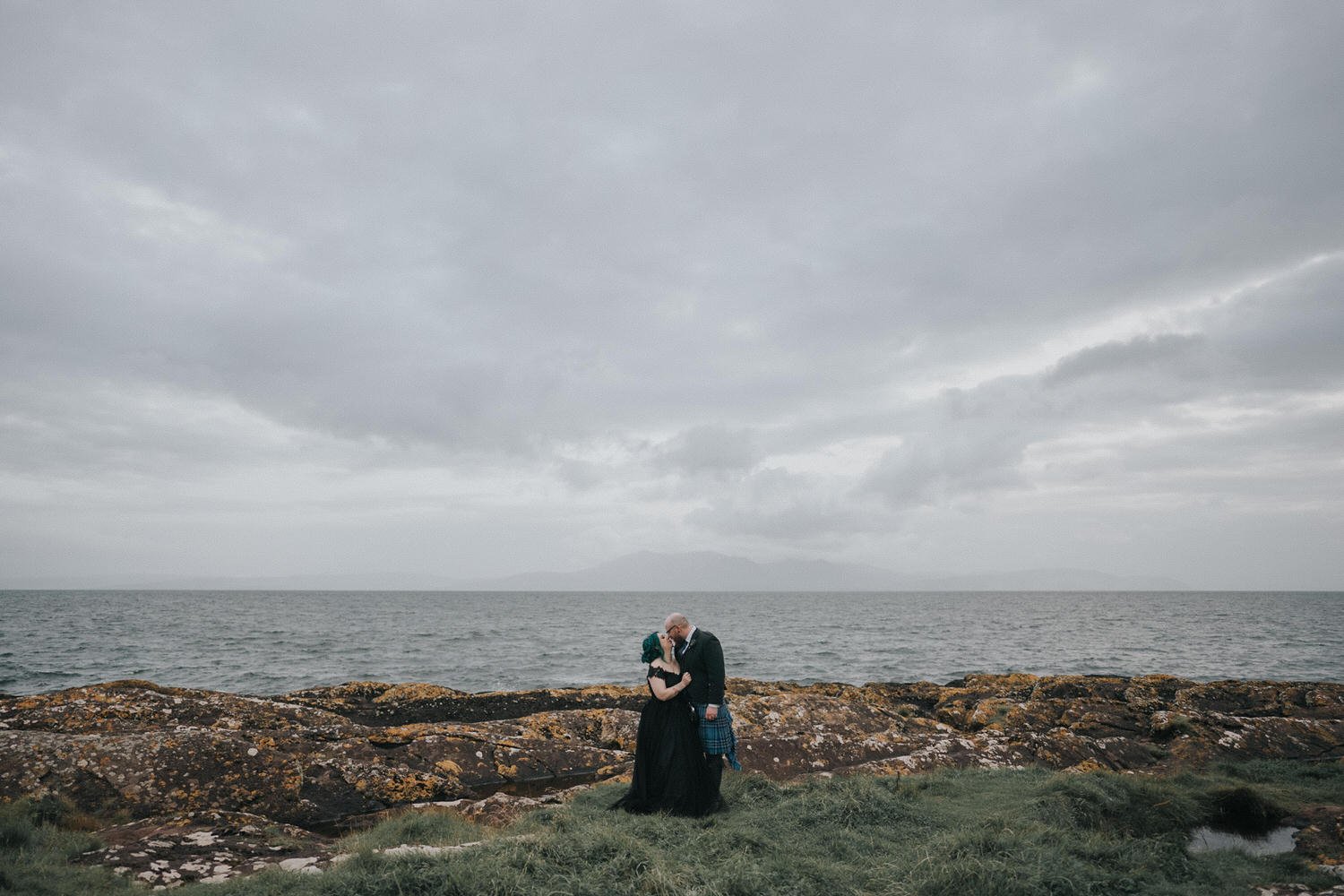 A couple share an intimate embrace on a rugged Scottish coastline, dressed in dark wedding attire with the sea and overcast sky stretching behind them, captured in a moody, documentary-style wedding photograph by Good Luck Wolf.