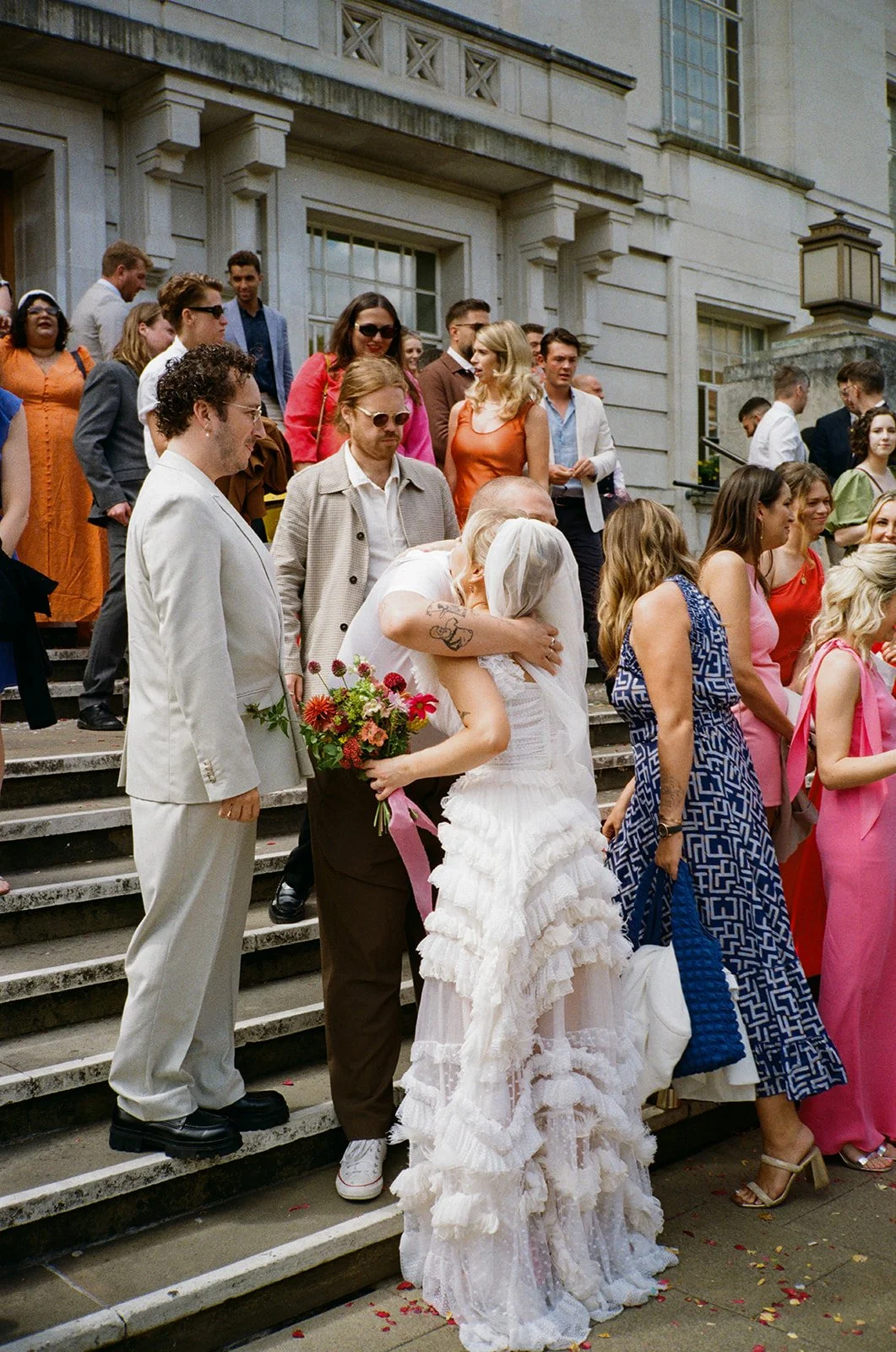 A bride in a tiered ruffled white gown and veil hugs a guest on the steps outside a wedding venue while holding a bouquet of bright flowers, surrounded by a large group of guests in colourful outfits.