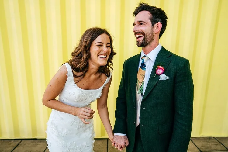 A newly married couple share a big laugh as they hold hands in front of a bright yellow backdrop, captured with warmth and character by Jordanna Marston Photography. Their relaxed expressions, bold colours and playful energy create a modern, upliftin