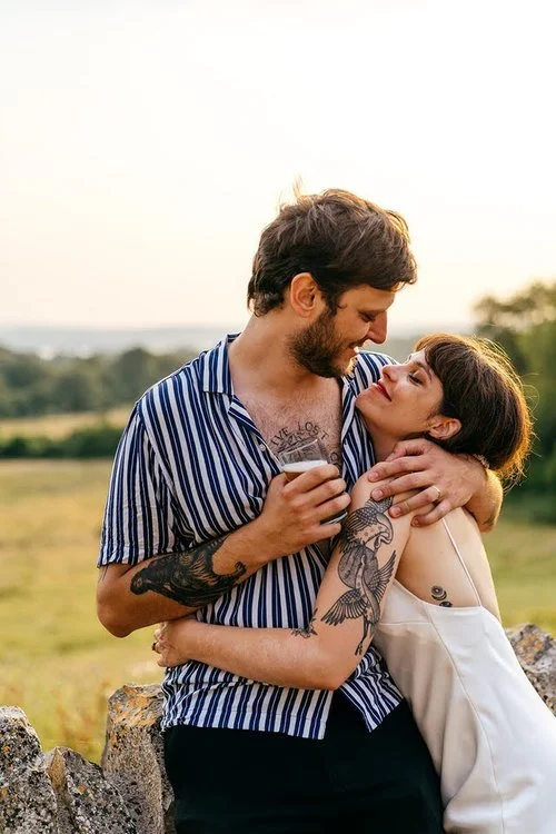 A couple share a warm, intimate embrace at sunset overlooking open countryside, beautifully captured by Jordanna Marston Photography. Their relaxed styling, meaningful connection and the soft golden light create a natural and heartfelt scene full of 