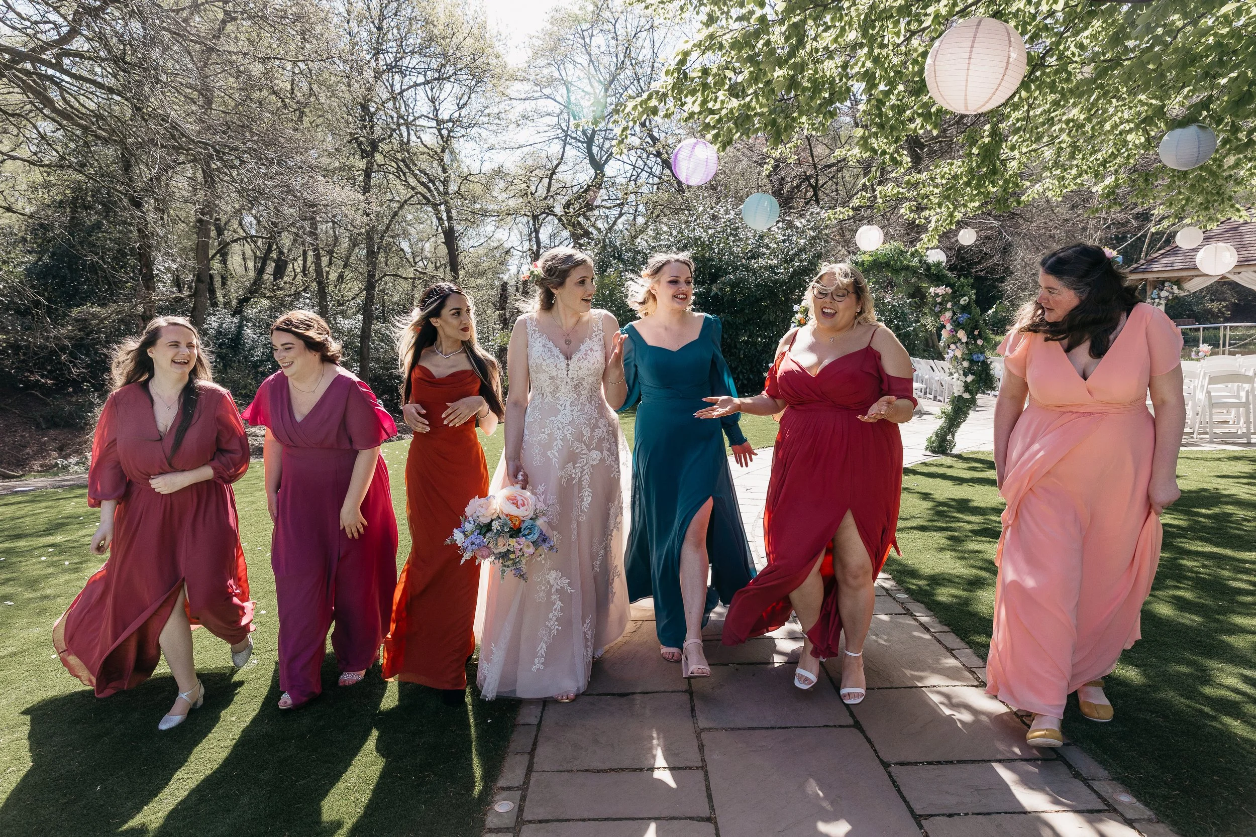 A bright and joy filled moment captured by YU Photography, showing the bride walking arm in arm with her bridesmaids beneath sunlit trees and hanging lanterns. Each bridesmaid wears a vibrant dress in shades of pink, red and teal, creating a colourfu