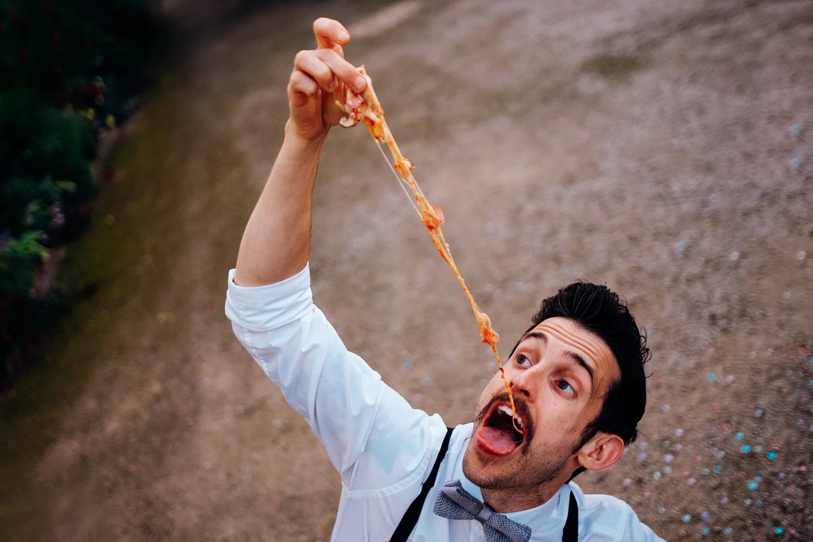 A man dressed in a white shirt with rolled sleeves, suspenders and a bow tie is pictured outdoors lifting a long, stretchy piece of melted cheese toward his mouth while looking up with an animated expression. The ground around him is scattered with s