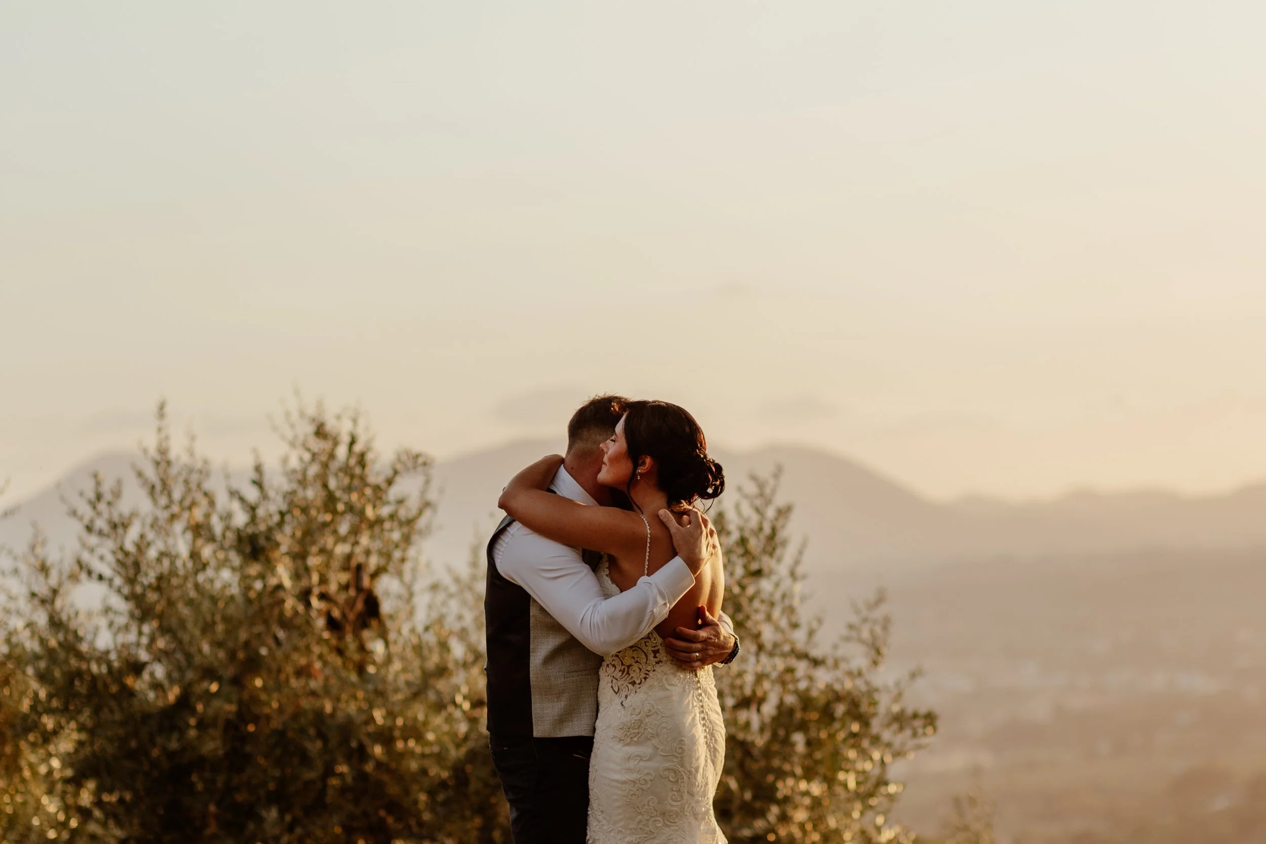 A warm and intimate golden hour wedding portrait captured by Laura Wilson Photography, showing a couple embracing against a soft sunset backdrop. The bride in a lace gown wraps her arms around her partner as they hold each other close, surrounded by 