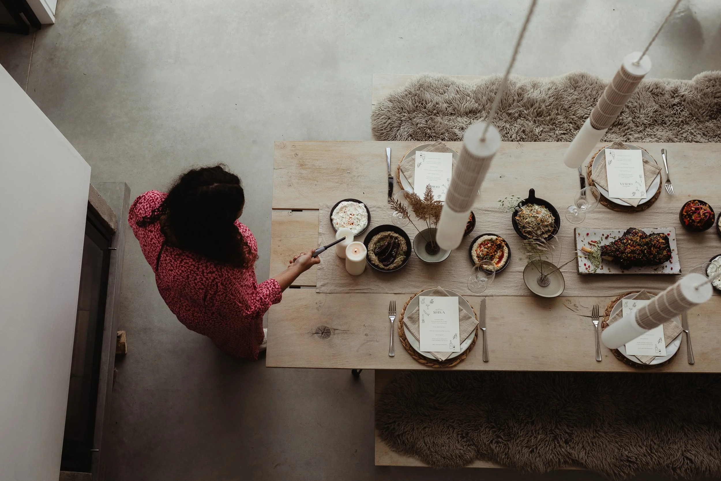 Overhead shot of a beautifully styled modern dining table set for an intimate gathering, featuring natural wooden textures, neutral linens, elegant menu cards and vibrant sharing dishes, as a woman in a pink patterned dress lights a candle to complet