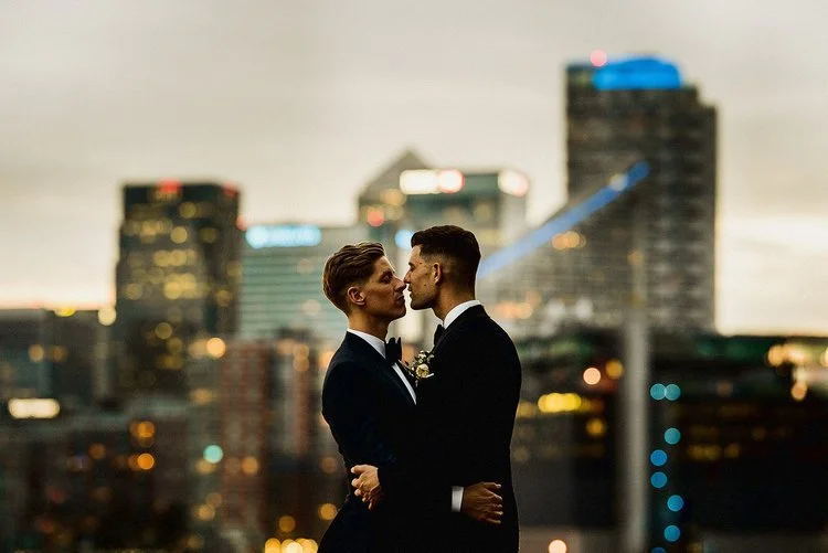 Two grooms stand close together on a rooftop at dusk, holding each other against a backdrop of glowing city lights. They lean in for a kiss, framed by tall buildings softened by evening haze. Their dark suits look sharp and modern, and the gentle way