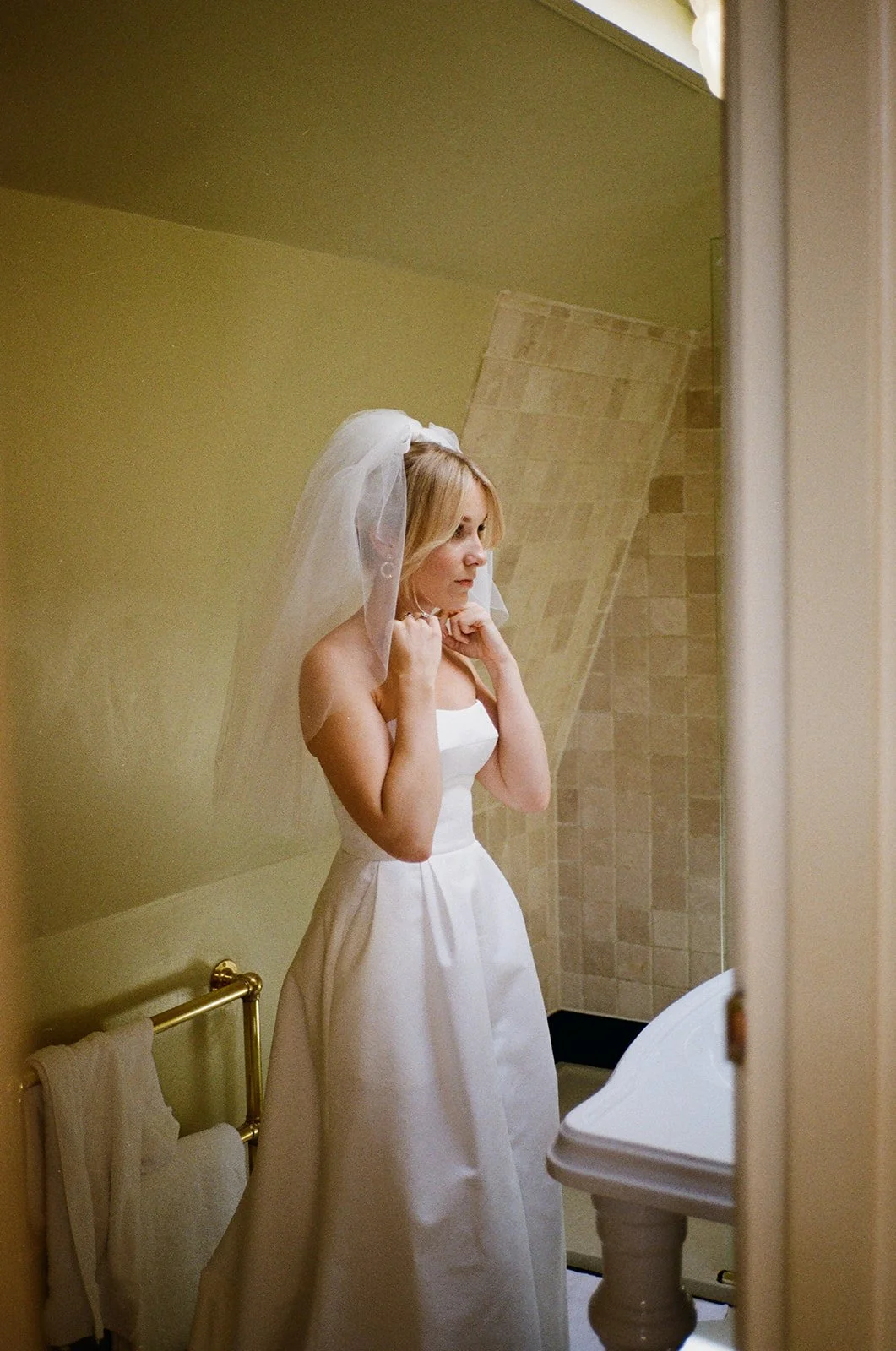 The bride stands quietly in a softly lit bathroom adjusting her necklace while wearing a simple strapless white gown and a short veil, her reflection partially visible in the mirror as warm light falls across the tiled wall behind her and the folded 