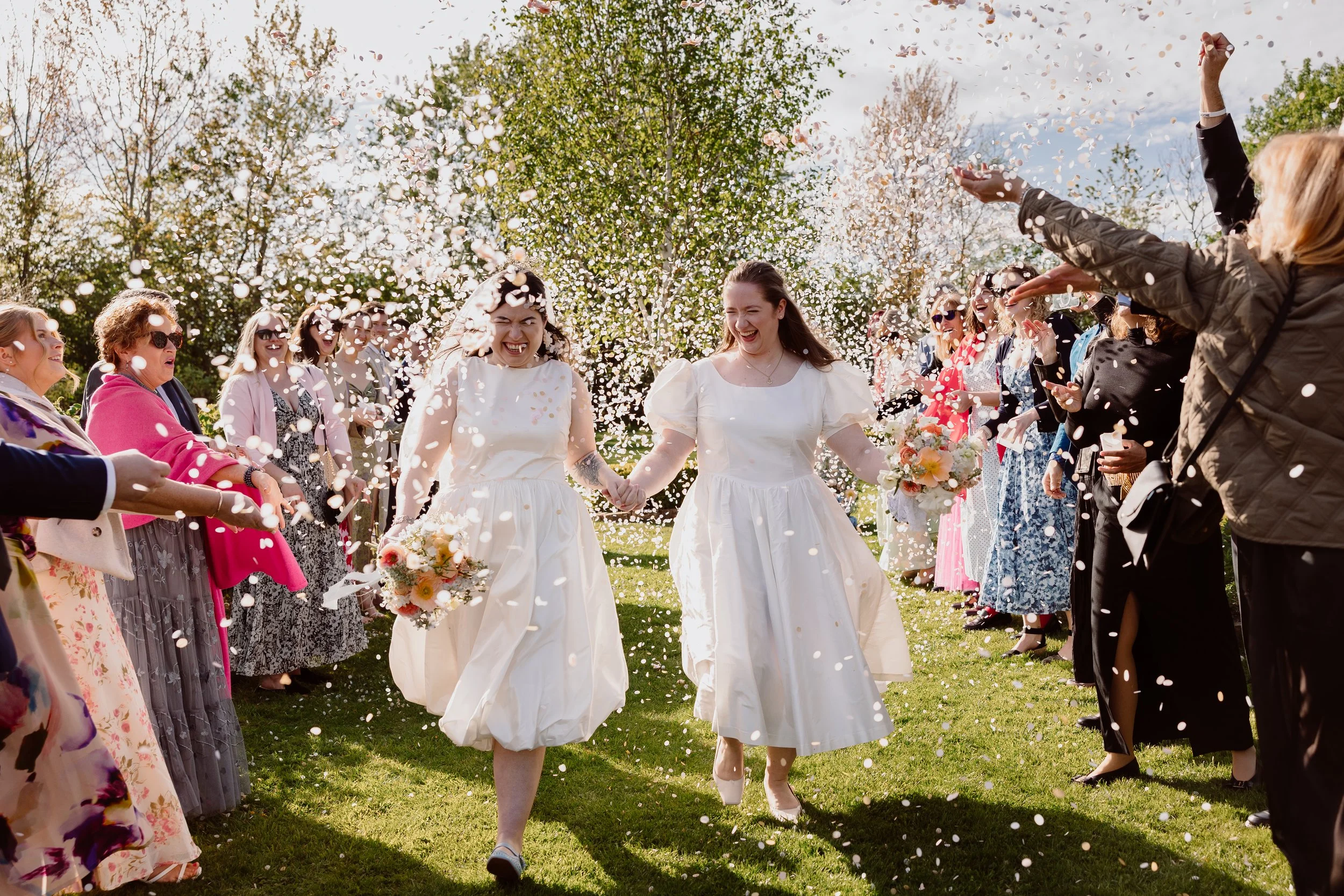 A joyful outdoor confetti moment captured by Joy Louise Photography, showing two brides walking hand in hand through a tunnel of cheering guests. Both brides wear elegant short white dresses and hold soft spring bouquets as they laugh together beneat