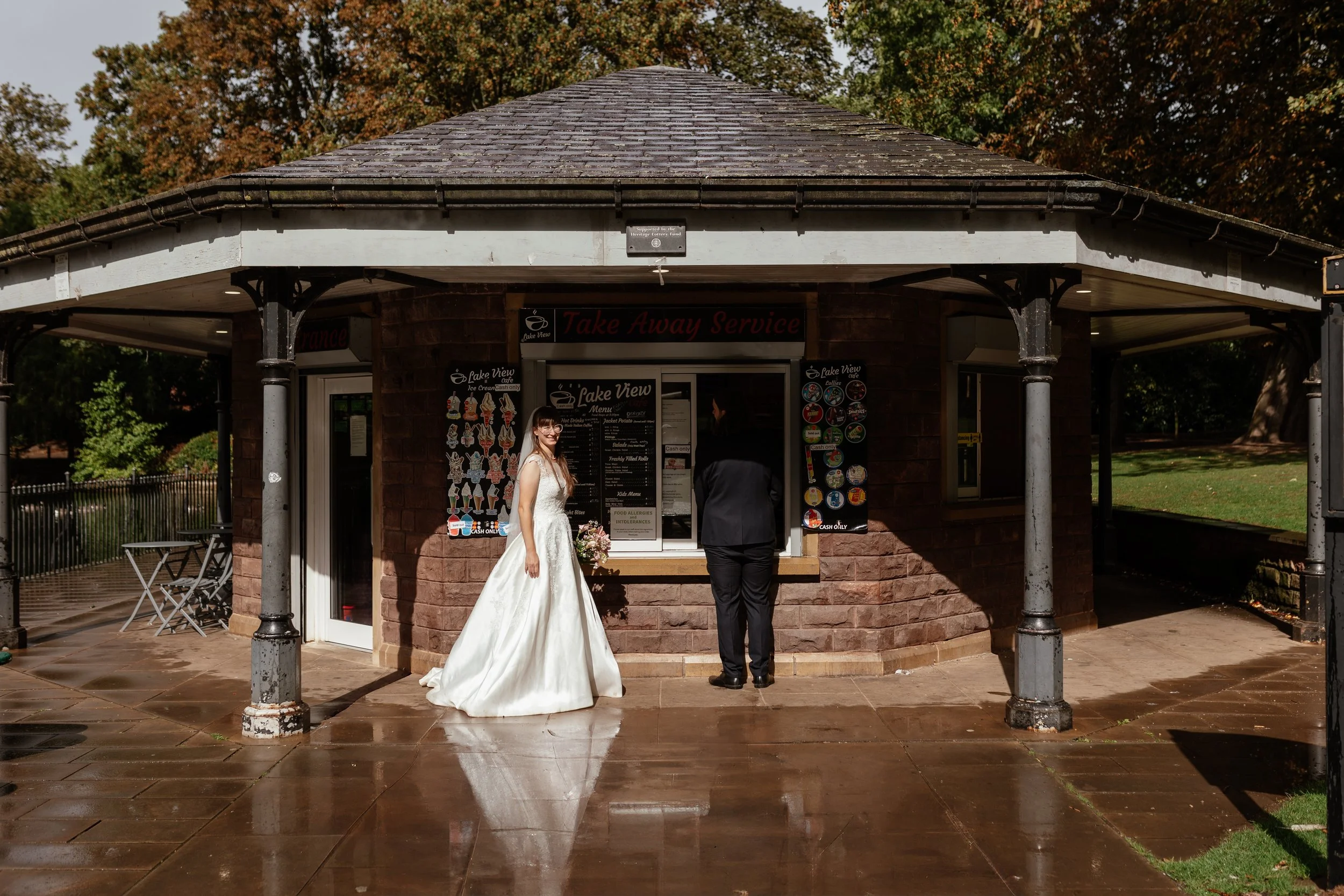 A playful and relaxed wedding moment captured by Laura Wilson Photography, showing a couple stopping at a park café for takeaway treats on their wedding day. The bride stands in a classic satin gown with floral detailing, smiling toward the camera wh