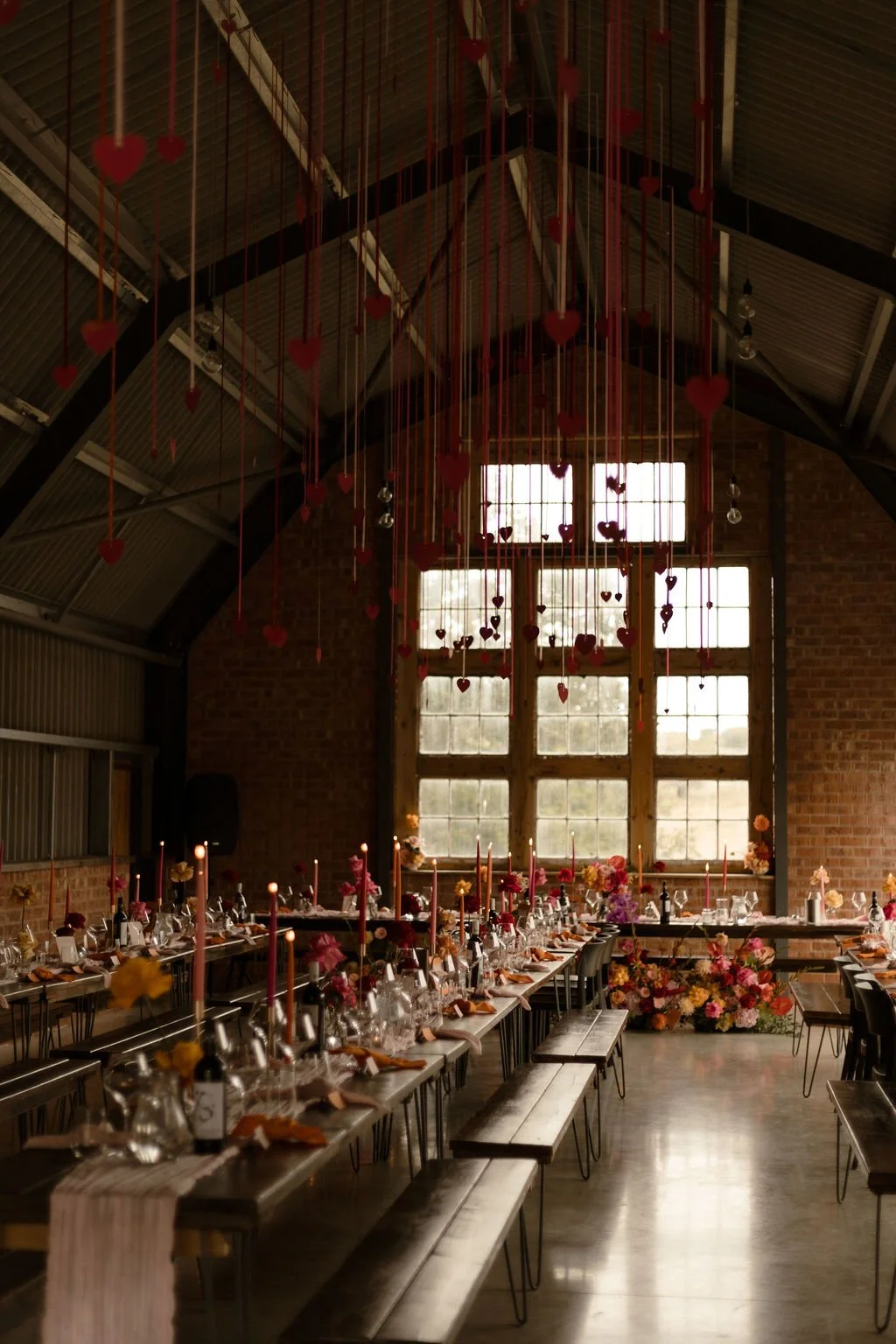 A romantic wedding reception setup inside The Giraffe Shed in Wales, featuring long banquet-style tables decorated with pink and orange floral arrangements, tall taper candles, and modern tableware. Red heart decorations hang elegantly from the indus