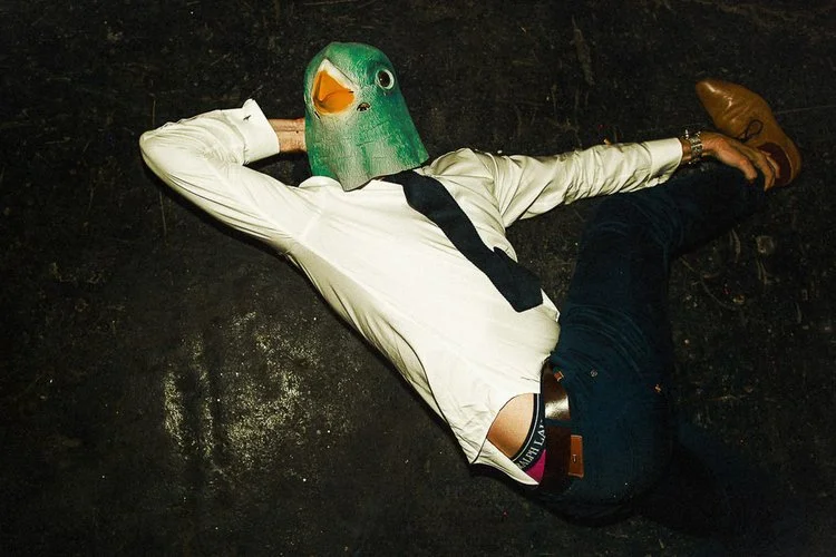 A person in formal wedding attire is lying sprawled across a dark floor, one arm bent behind their head and the other stretched outward. They’re wearing a white shirt, dark trousers, and dress shoes, with part of their waistband showing as their shir