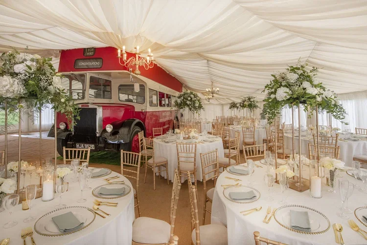 A beautifully styled wedding marquee featuring the iconic red Deckerdence double-decker bus bar as the stunning centerpiece. The elegant setup includes round tables with gold chairs, crystal glassware, and soft white floral arrangements, creating a l
