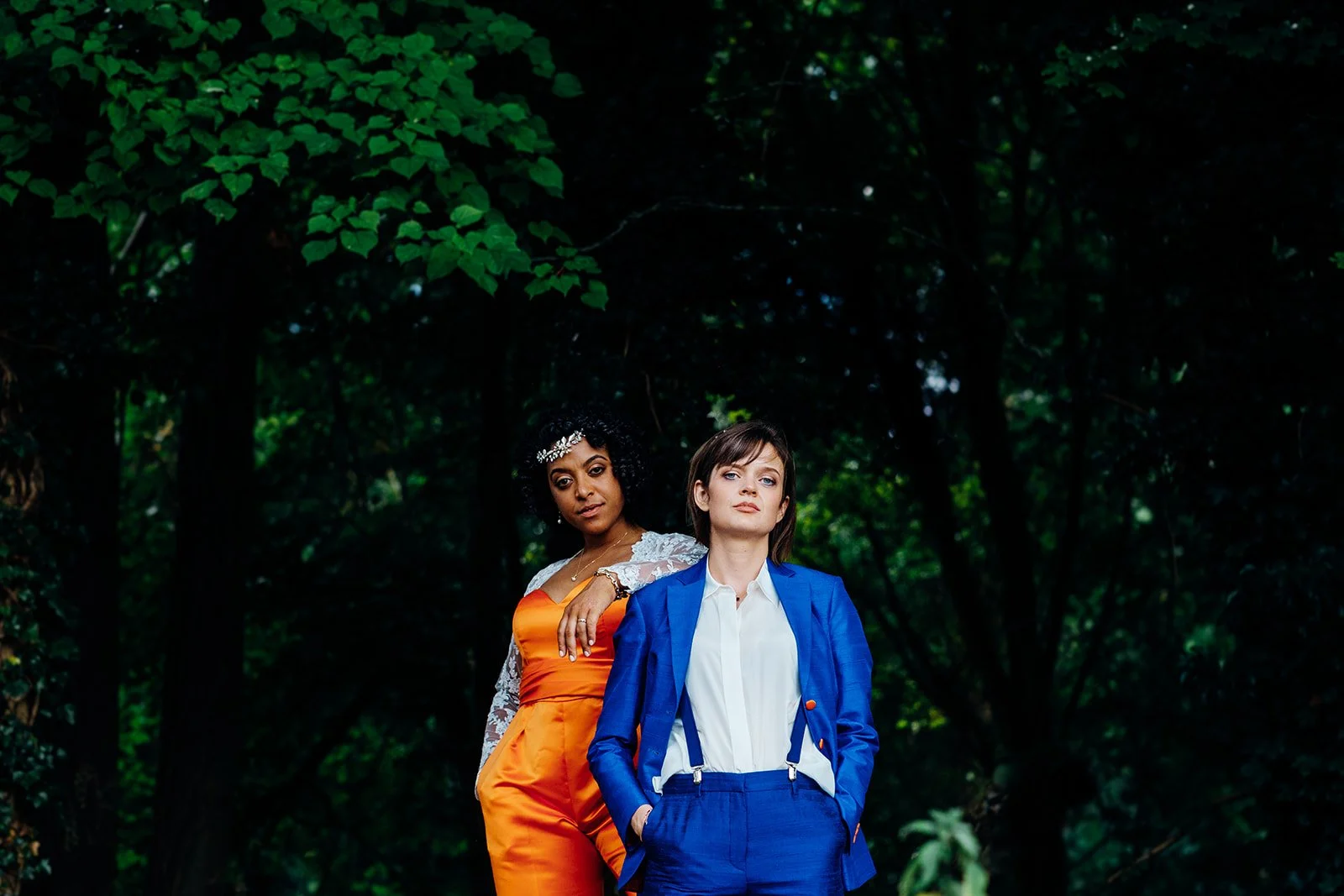 Two women stand side by side in a wooded outdoor setting, framed by deep green foliage, with one wearing a bright orange outfit with lace sleeves and the other wearing a tailored blue suit with a white shirt and suspenders. Both look directly toward 