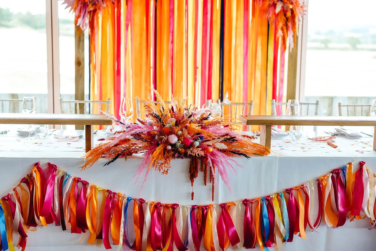 A vibrant and celebratory tablescape filled with colour, featuring a large dried flower arrangement in vivid shades of hot pink, orange, red, coral and touches of blue, with feathery textures and soft pompom-like accents spreading wide across the cen