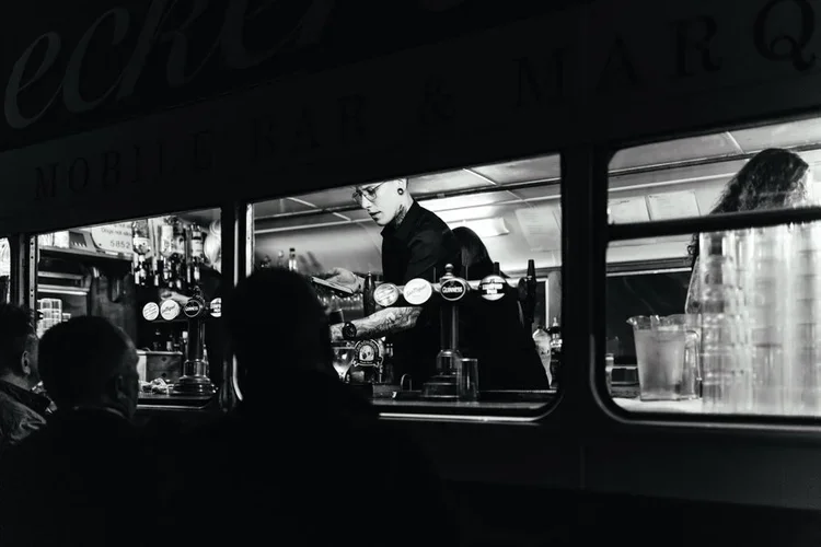 A black-and-white photo capturing the stylish Deckerdence Mobile Bar in action, housed inside a converted vintage double-decker bus. The bartender serves drinks through the open window to guests outside, creating an intimate and atmospheric moment. T