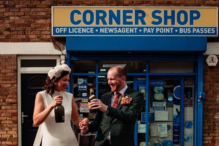A fun and energetic wedding moment captured by Natalia Case Photography, showing the couple celebrating outside a corner shop with bottles in hand. The bright signage and urban backdrop create a playful contrast to their wedding attire, giving the sc