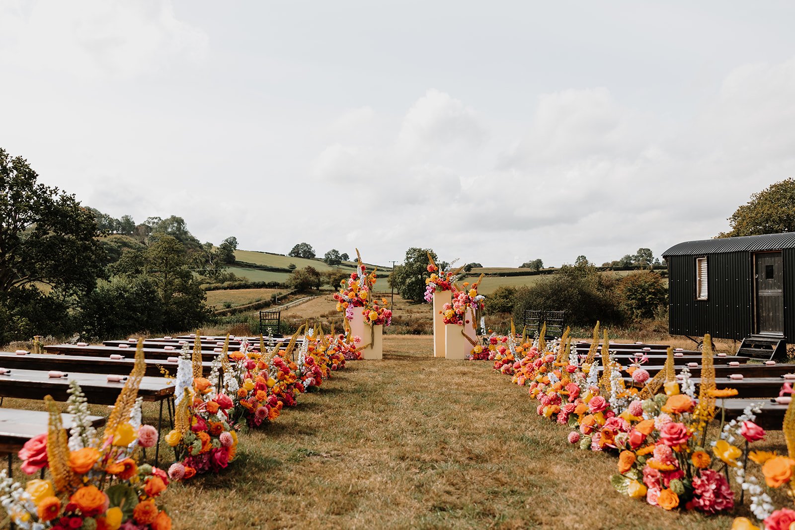 A stunning outdoor wedding ceremony setup at The Giraffe Shed in Wales, featuring a vibrant floral aisle lined with bold pink, orange, and yellow blooms. The ceremony backdrop showcases modern pastel arches adorned with colourful flowers, perfectly f